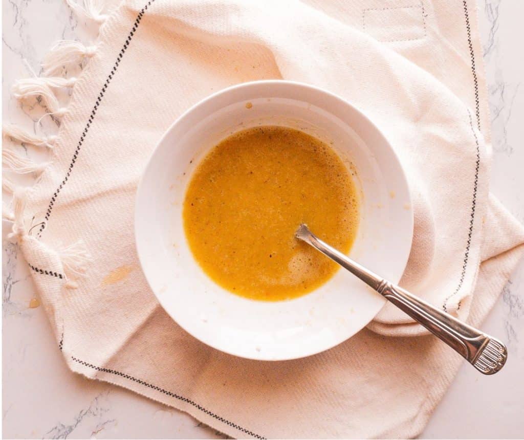 a yellow colored honey salad dressing in a white bowl with a spoon and kitchen towel underneath it.