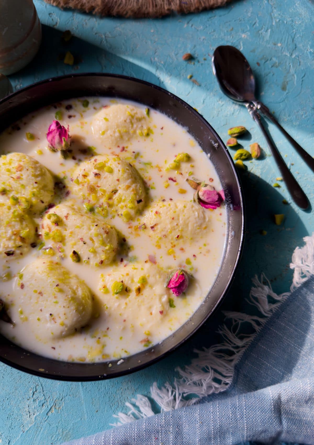 garnished rasmalai served in black deep plate with spoons on the side.
