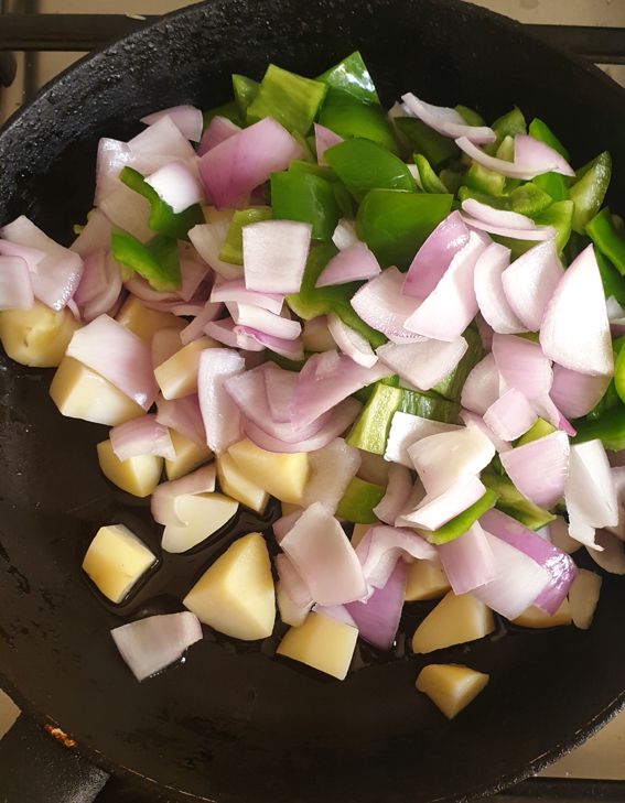 raw cube cut onions, potatoes and capsicum in a black frying pan. 