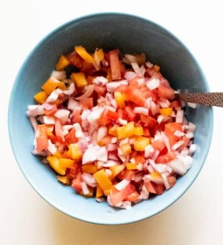 finely chopped onions, tomatoes and orange bell pepper in a blue bowl with a spoon in it