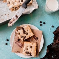 2 pieces of blonde chocolate chip brownie with a glass of milk on the side and a half showing tray of chocolate chip brownies with a few scattered chocolate chips all around a blue backdrop