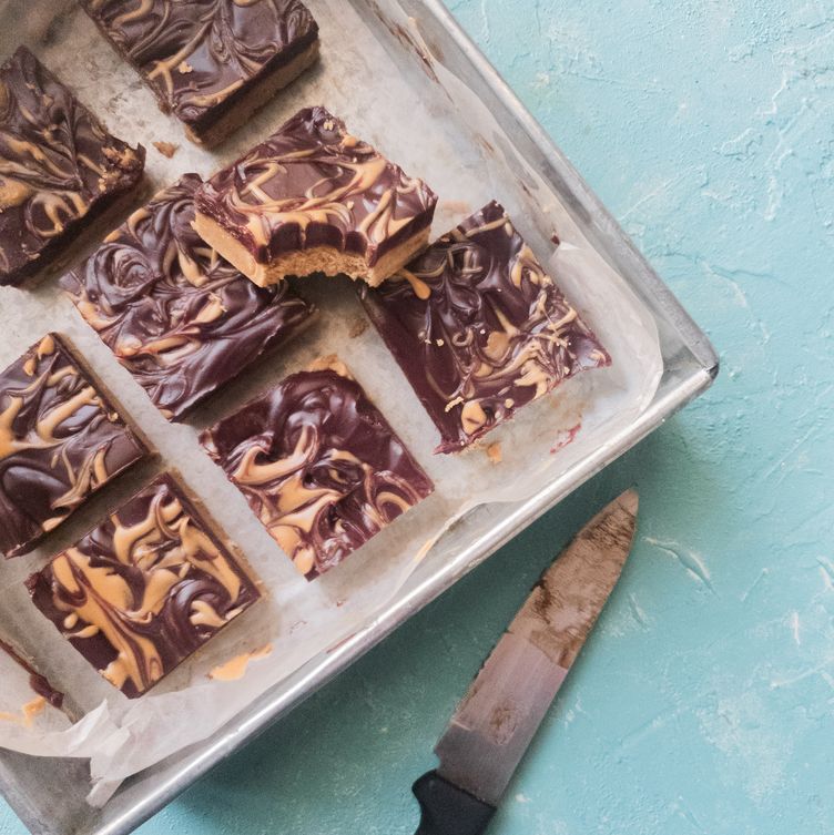 a partial view of a square baking tray lined with parchment paper. It has a few chocolate peanut butters bars on it and one piece with a bite taken. there is a partial view of a knife that has been used to cut the bars. the background is light blue