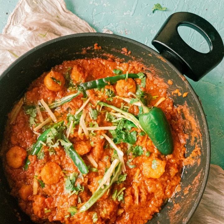 a top view picture of prawn masala garnished with green chilies and ginger in a black wok, kept on top of a cream napkin against a light blue blackdrop