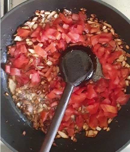 tomatoes added in a pot of golden brown onions, with a wooden spoon on top