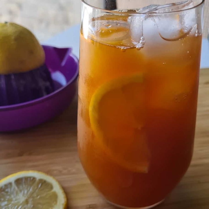 a glass of orange hued lemon iced tea with slices of lemon and ice cubes visible in the glass