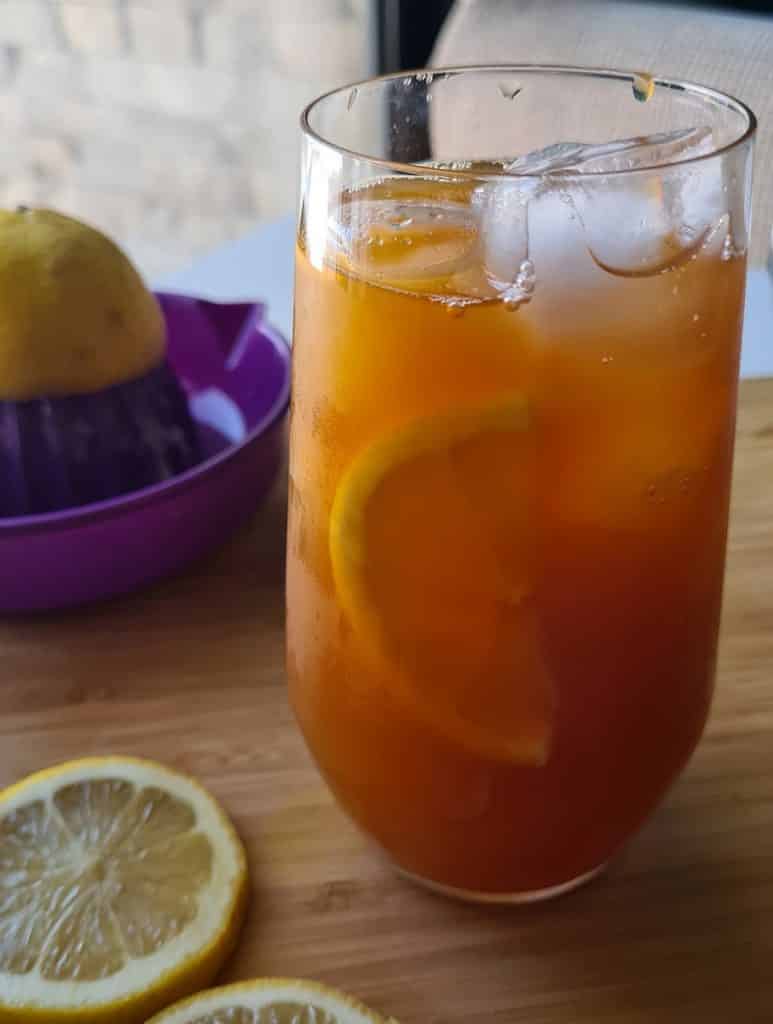 a glass of orange hued lemon iced tea with slices of lemon and ice cubes visible in the glass