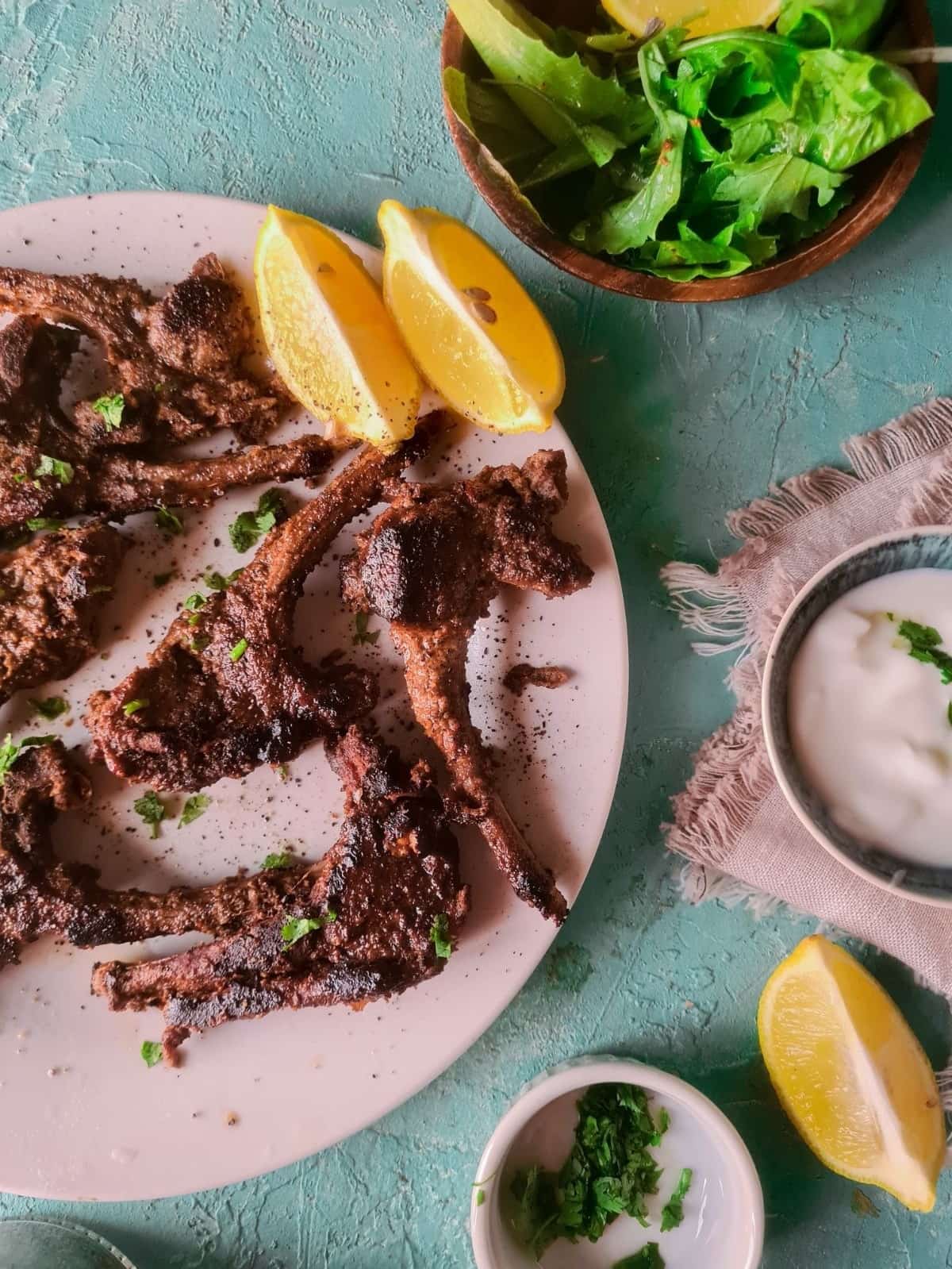 Grilled and steamed mutton chops ready on a white plate with lemon slices on the side and a bowl with yogurt.